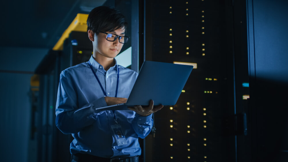 Male IT Specialist Stands Beside the Row of Operational Server Racks, Uses Laptop for Maintenance.