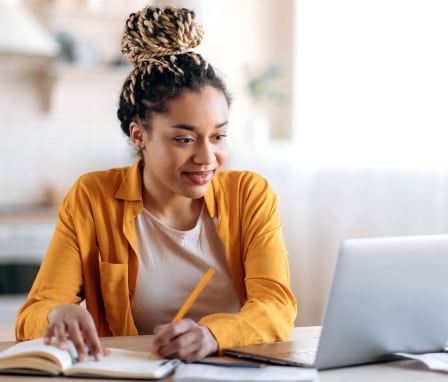 Woman writing in notebook while looking at laptop computer screen